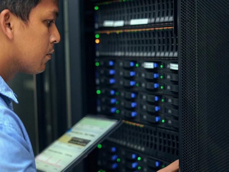 A man, using a mobile tablet like a clipboard, looks over the numerous brightly colored lights on a server.