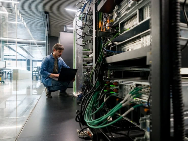 Another man crouches near the floor as he inspects the cables and other elements in a wall of technology equipment.